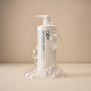 A white pump bottle of O&M Original Detox Shampoo standing on a surface covered with clear bubble-like foam, against a warm beige studio background.